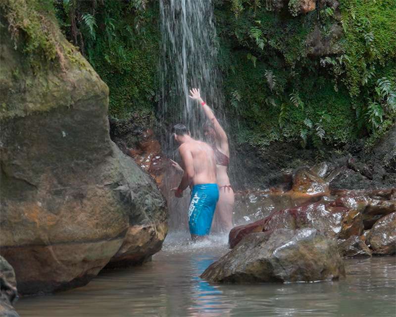Man standing under the waterfall at Caldeira Velha, dark volcanic rock and ferns behind him
