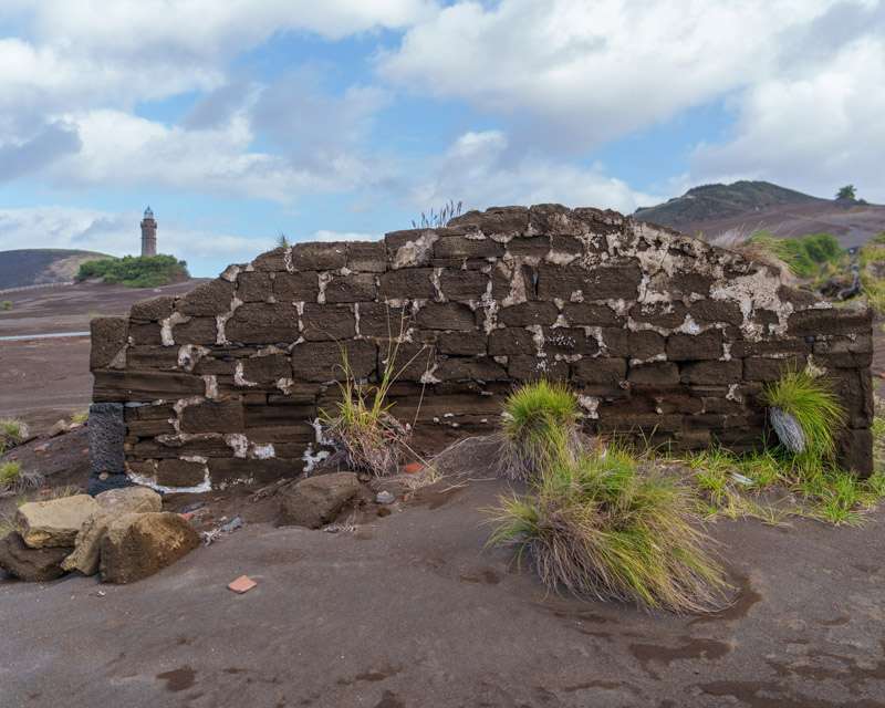 Dry-stone ruins standing in the grass with the Capelinhos lighthouse on the ridge behind