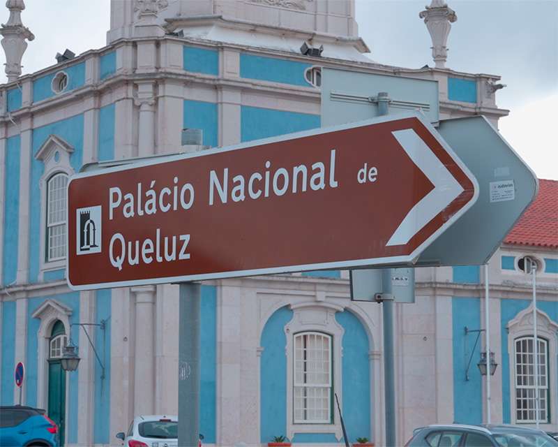 Braunes Hinweisschild zum Palácio Nacional de Queluz vor der blau-weißen Palastfassade