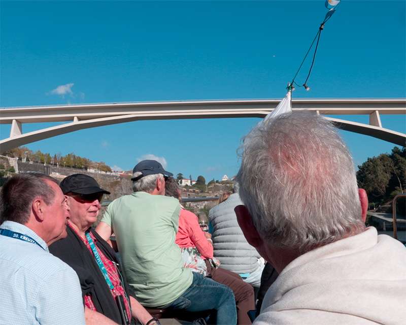 Passengers seated on the open deck of a Douro tour boat passing under a white concrete arch