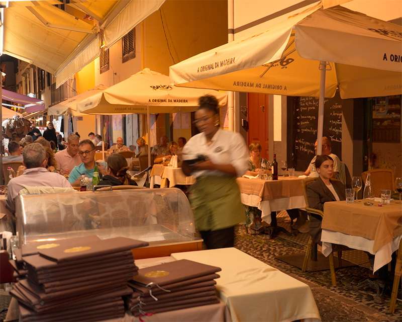 Busy restaurant terrace with white parasols and a waiter on Rua de Santa Maria, Funchal, in the evening