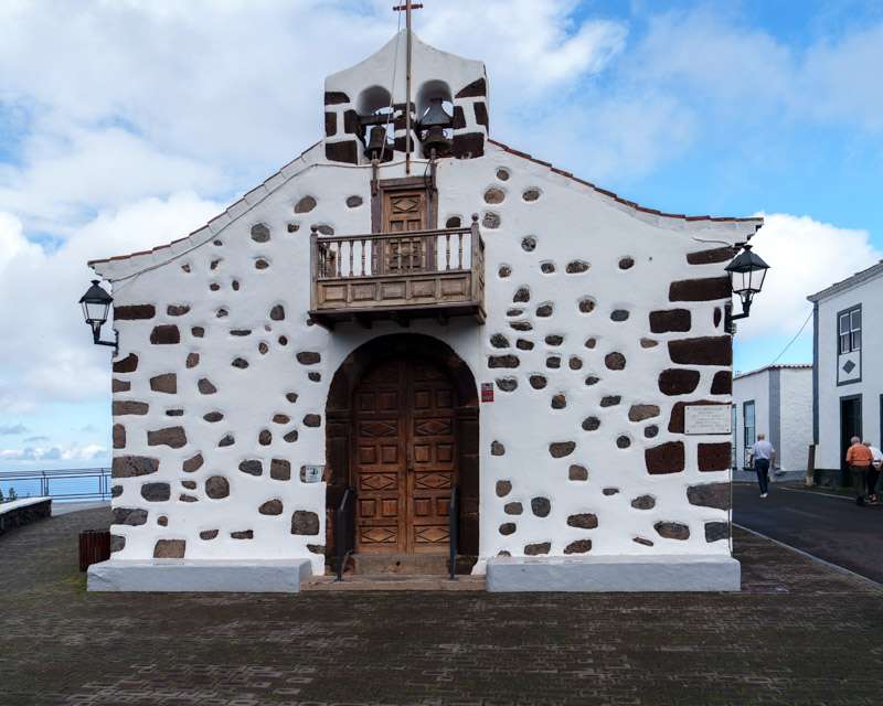 Whitewashed chapel at San Bartolomé with black volcanic stonework framing the door and a wooden bell gable