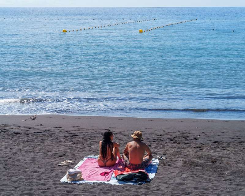 Two people sitting on the dark sand behind the buoyed swimming area at Puerto de Tazacorte