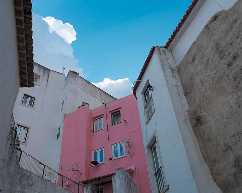 Schmale Gasse in der Alfama zwischen einem rosa Haus und einer weißen Wand unter blauem Himmel