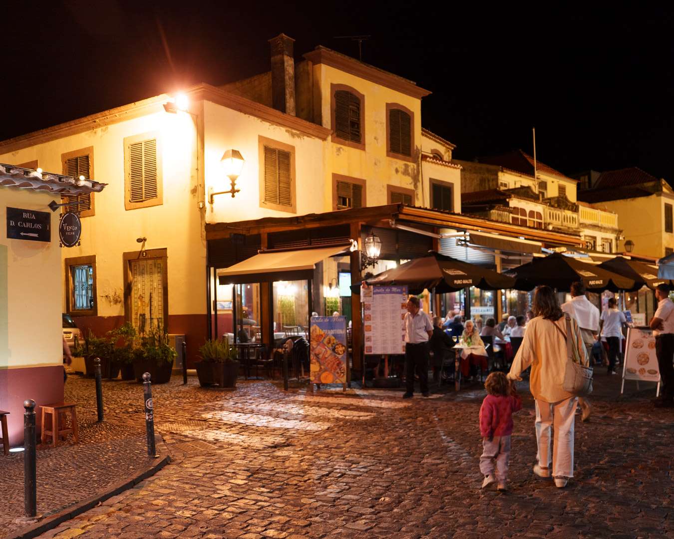 People walking the cobbled Rua de Santa Maria in Funchal old town in the evening, lit terraces on both sides
